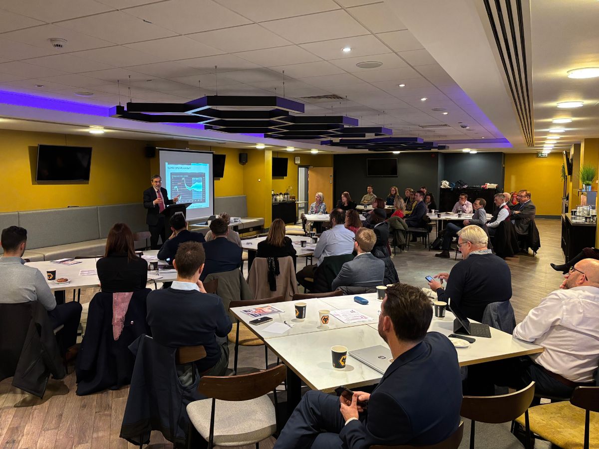 Image of Bank of England event hosted at Wolves Molineux Stadium, in the conference room. There are 6 tables with people sat around with drinks and paper. Listening to Graeme Chaplin presenting in front of a projector screen at the front of the room.